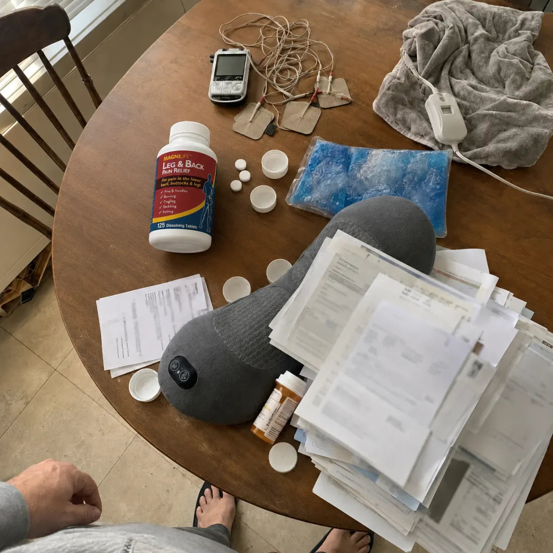 A table covered with failed remedies: the massage pillow, pain-relief pills, wired electrode pads, a heating pad, an ice pack, prescription bottles, and a tall stack of medical bills