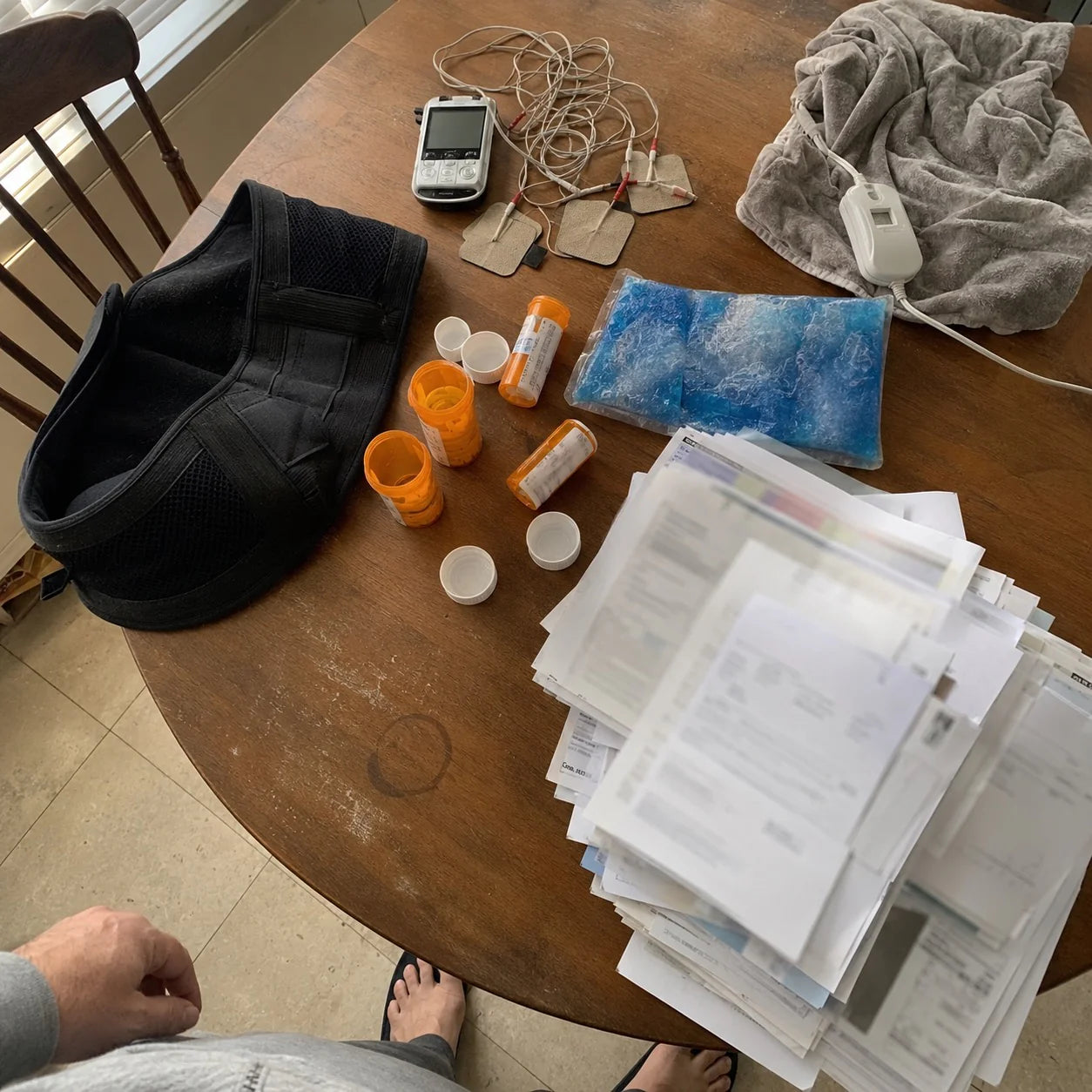 A pile of failed remedies on a kitchen table: pills, a back brace, heat, ice, a wired pad, and a stack of bills